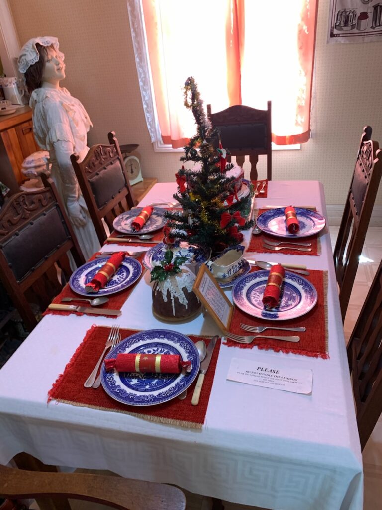 A table in an 1860s kitchen with a white tablecloth set for Christmas dinner. There is a mannequin dressed in white standing behind the table. Red Christmas crackers sit on blue willow dinner plates on red placemats. There is a small Christmas tree on the table decorated with gold and silver tinsel and red bows. In the front is a Christmas pudding with cream dripping down the side and a sprig of holly on top.