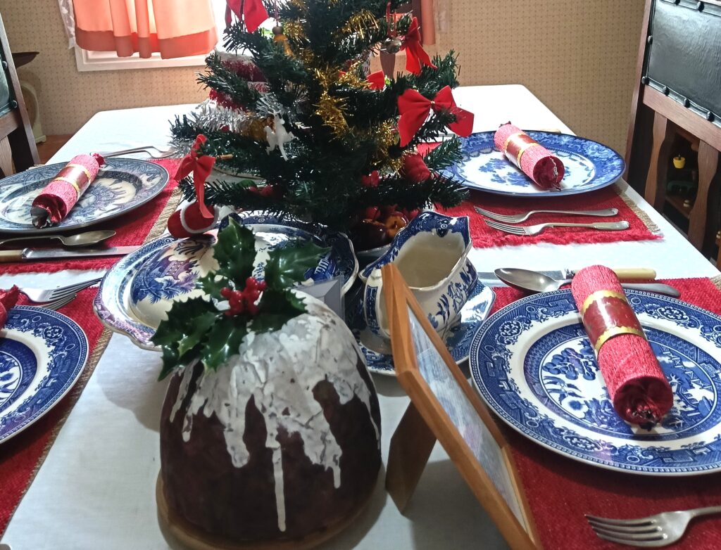 A table with a white tablecloth set for Christmas dinner. Red Christmas crackers sit on blue willow dinner plates on red placemats. There is a small Christmas tree on the table decorated with gold and silver tinsel and red bows. In the front is a Christmas pudding with cream dripping down the side and a sprig of holly on top.